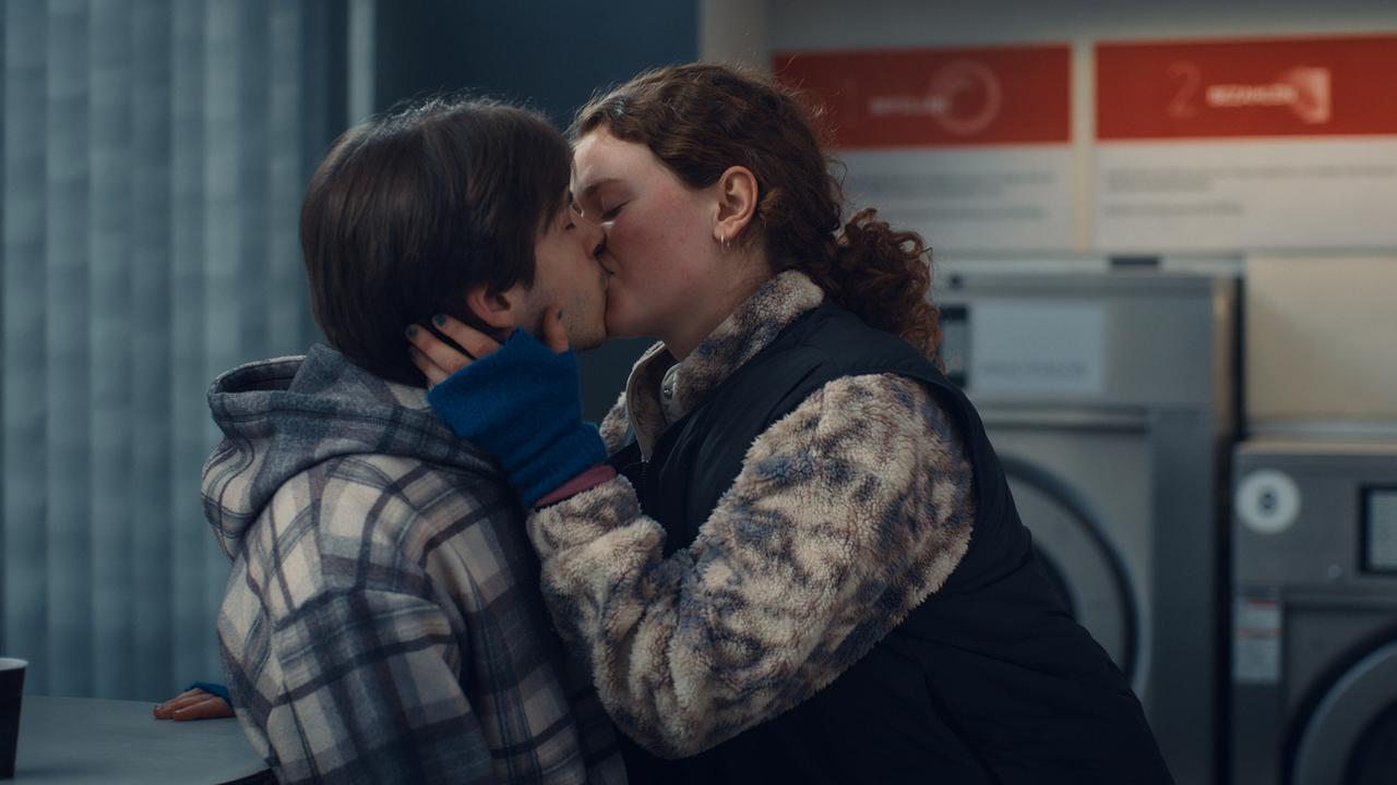 Luca Rossi und Fabienne küssen sich in einem Waschraum mit Waschmaschinen im Hintergrund.