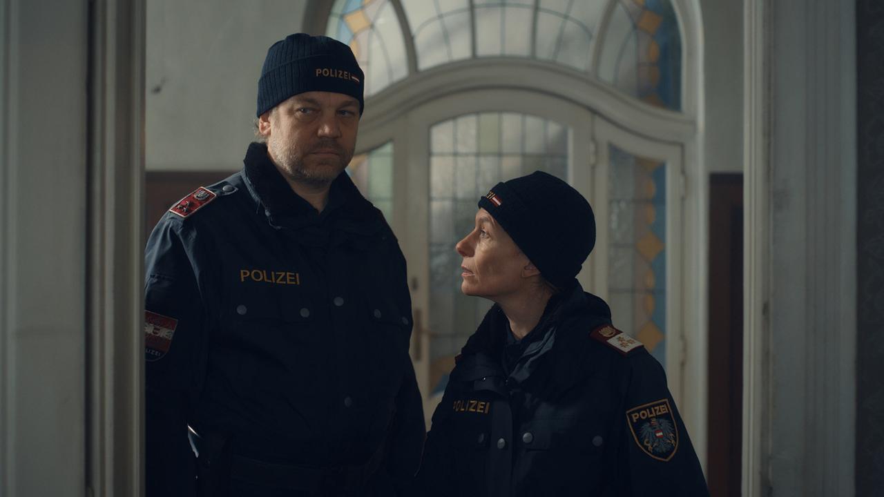 Polizist Günther und Polizistin Corina stehen in einem Gebäude mit großen verzierten Türen.