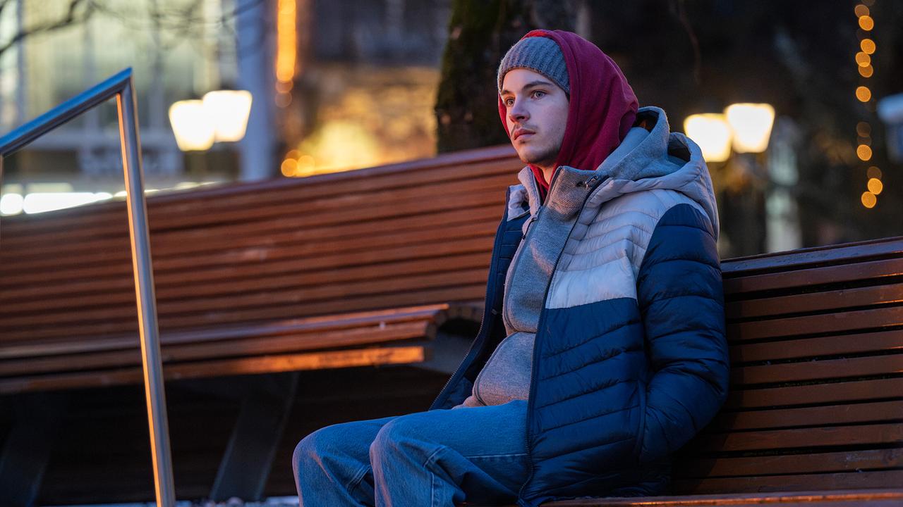 © ORF/BR/Superfilm/Stefanie Leo Luca Rossi sitzt abends warm angezogen auf einer Parkbank in einer beleuchteten Stadtumgebung.