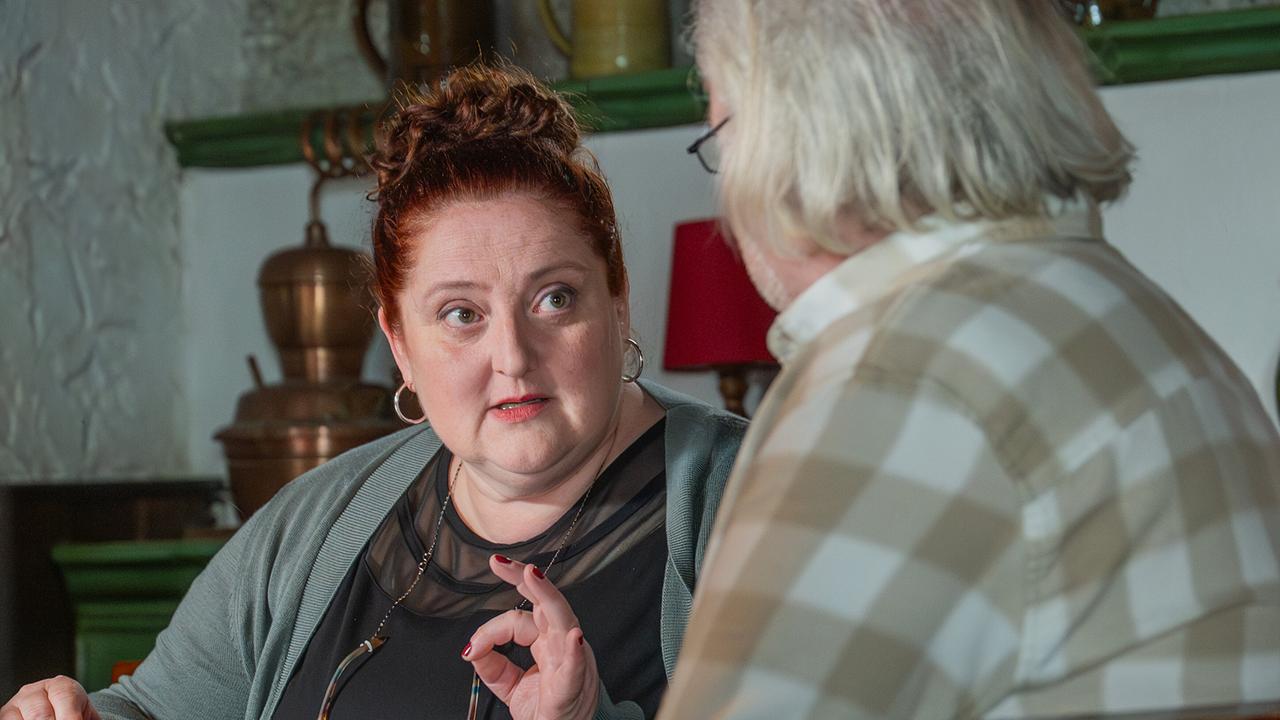 Petra und Schiesstl unterhalten sich in einer rustikalen Stube, umgeben von Dekoration und Holzmöbeln.