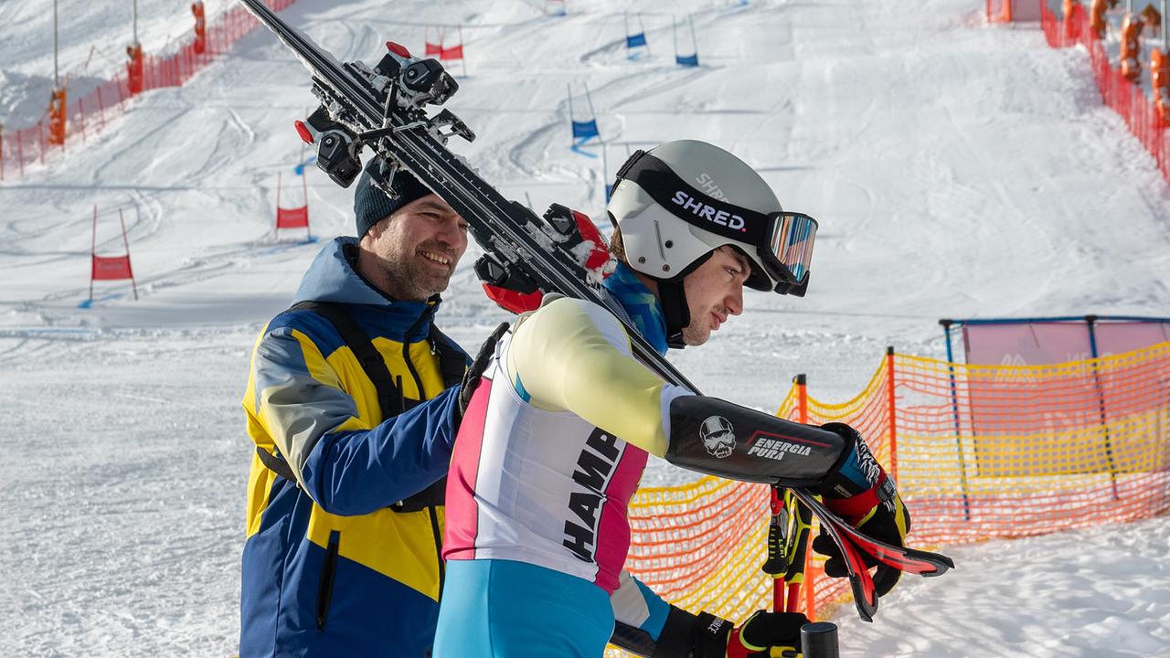 Mark Auer in Winterbekleidung legt seine Hand auf den Rücken von Nikki Auer im Skirennanzug und Helm mit den Skiern auf seinen Schultern, im Hintergrund eine schneebedeckte Piste.