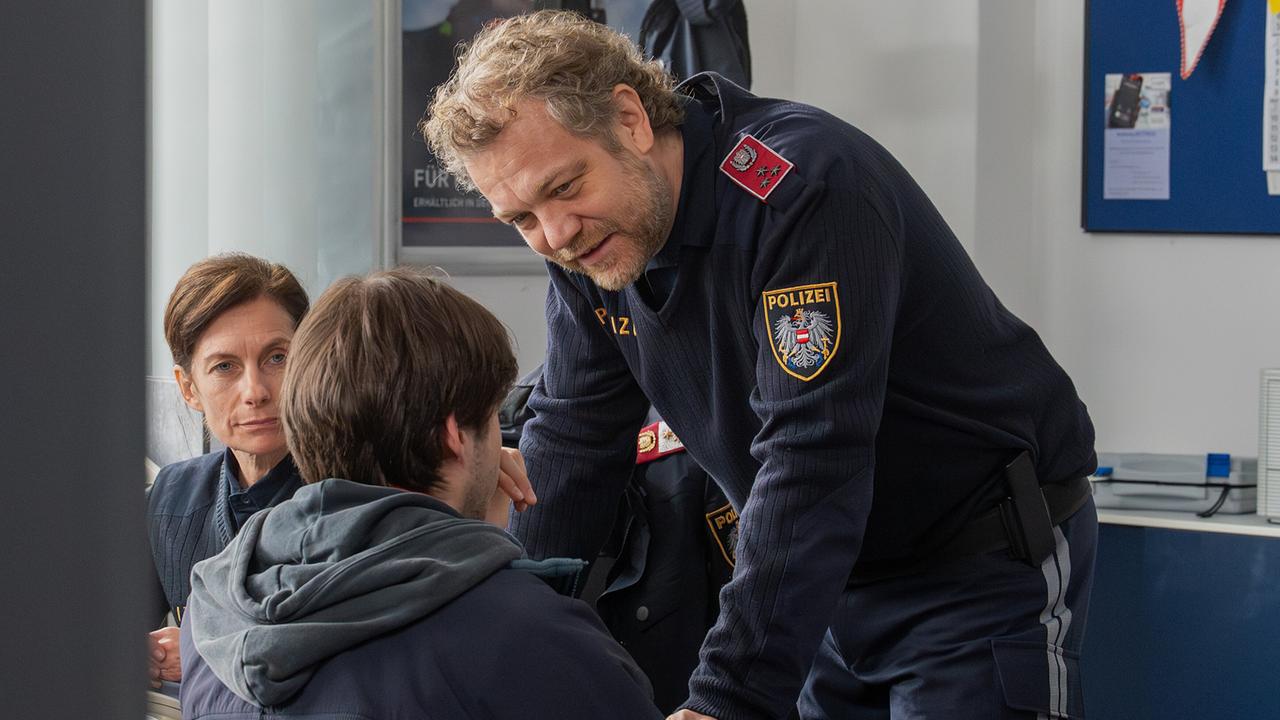 Polizist Günther beugt sich in einem Polizeibüro vor zu Luca Rossi, der auf einem Sessel sitzt. Polizistin Corina sieht im Hintergrund zu.
