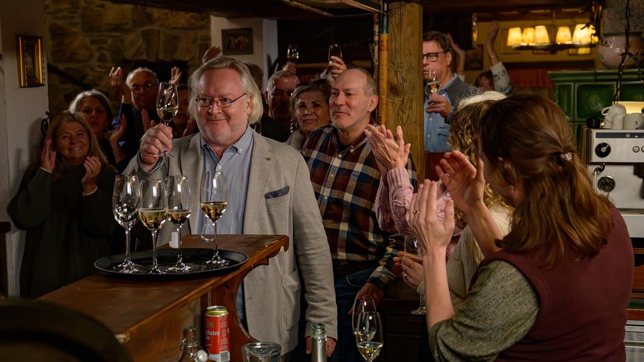 Schiesstl hebt ein Glas Wein an einer Bar in einem rustikalen Lokal, während die Umstehenden begeistert applaudieren.