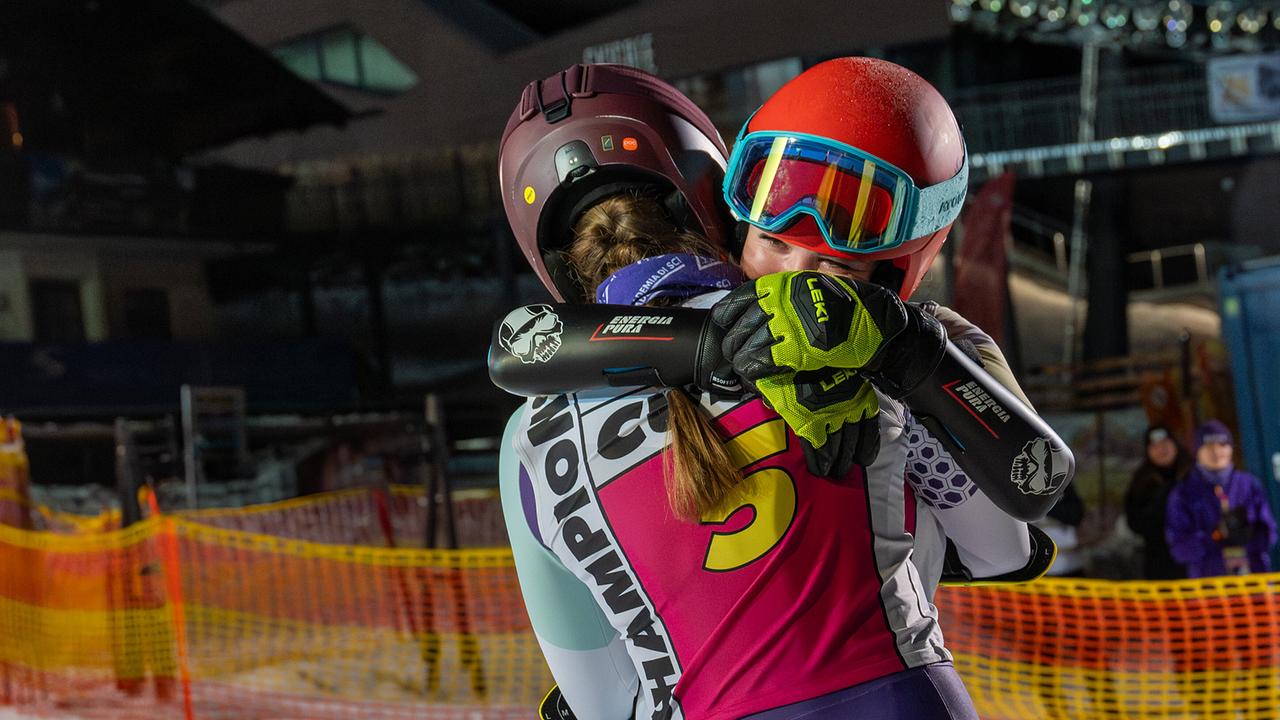 Die zwei Skirennläuferinnen Dani Strobl und Solenn Eden umarmen sich im Zielbereich eines Nachtrennens zwischen Absperrnetzen und Zuschauern.