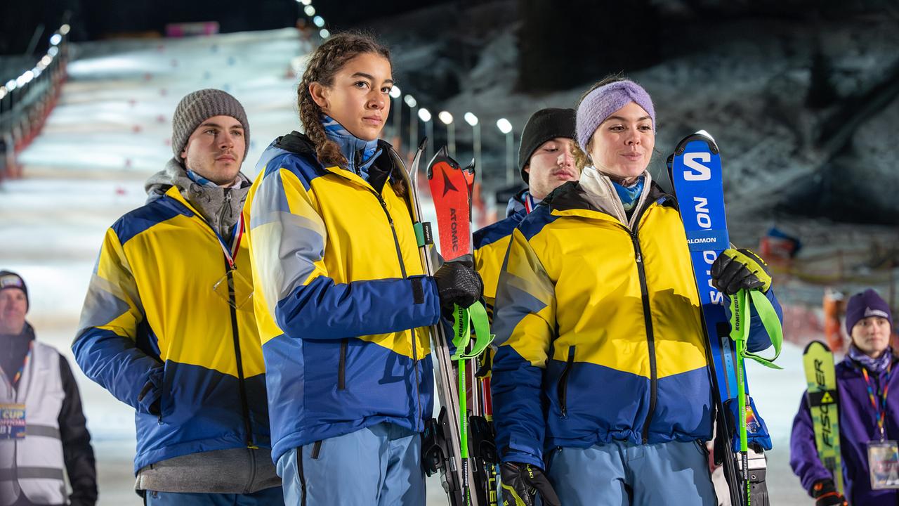 Luca Rossi, Nawal Ammann, Nikki Auer und Solenn Eden in einheitlicher gelb-blauer Kleidung stehen mit Skiern in der Hand am Rand einer nächtlichen Rennstrecke.