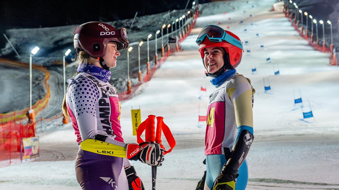 Dani Strobl und Solenn Eden im Skirenn-Outfit stehen sich unter Flutlicht am nächtlichen Hang gegenüber und führen ein Gespräch.