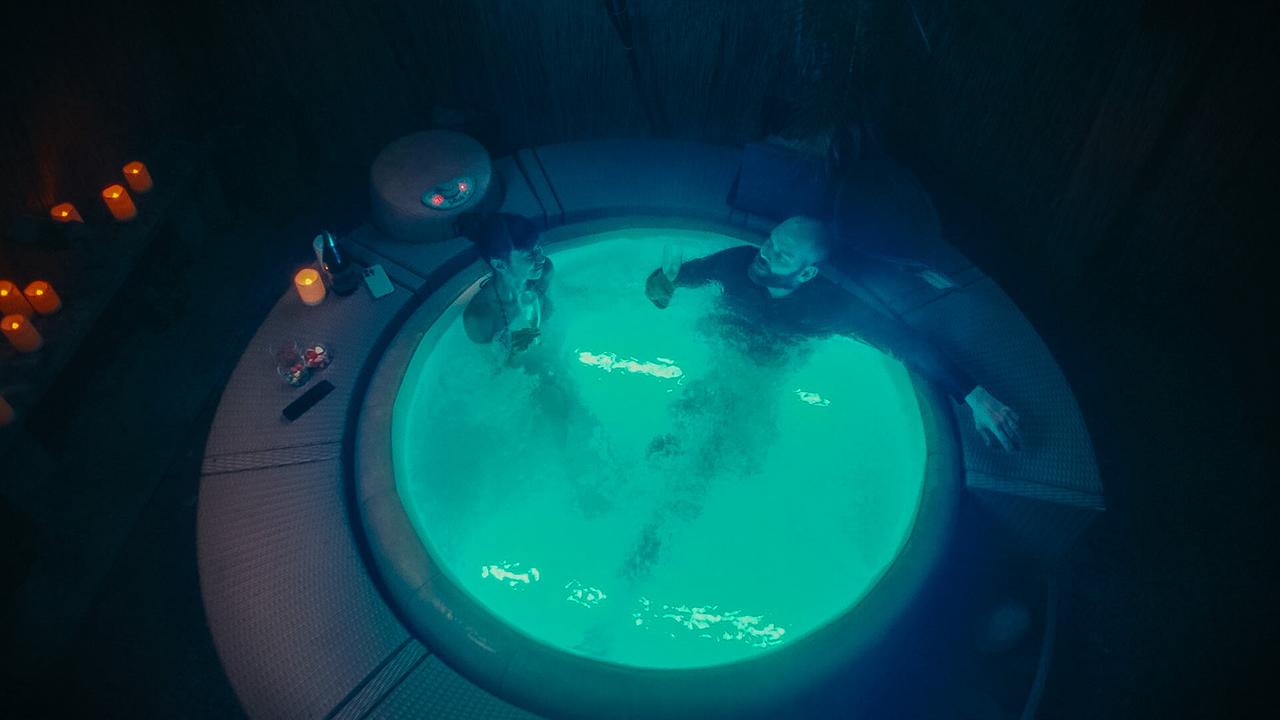 Cat Jiminez und Igor Karbus sitzen mit Sektgläsern bei Dunkelheit in einem runden, blau beleuchteten Whirlpool. Rund um den Whirlpool stehen Kerzen und Getränke.