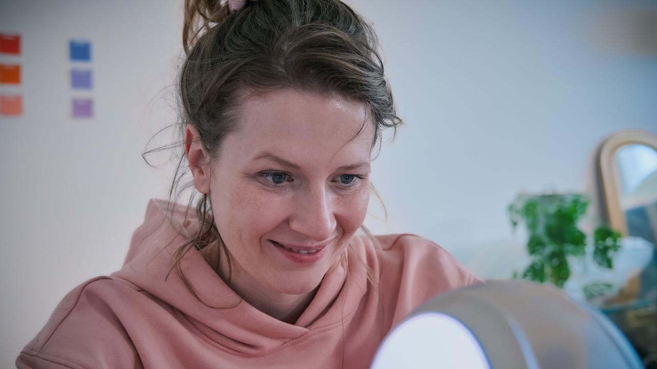 Anna in einem rosa Hoodie sitzt konzentriert an einem beleuchteten Gerät in einem hellen Raum.