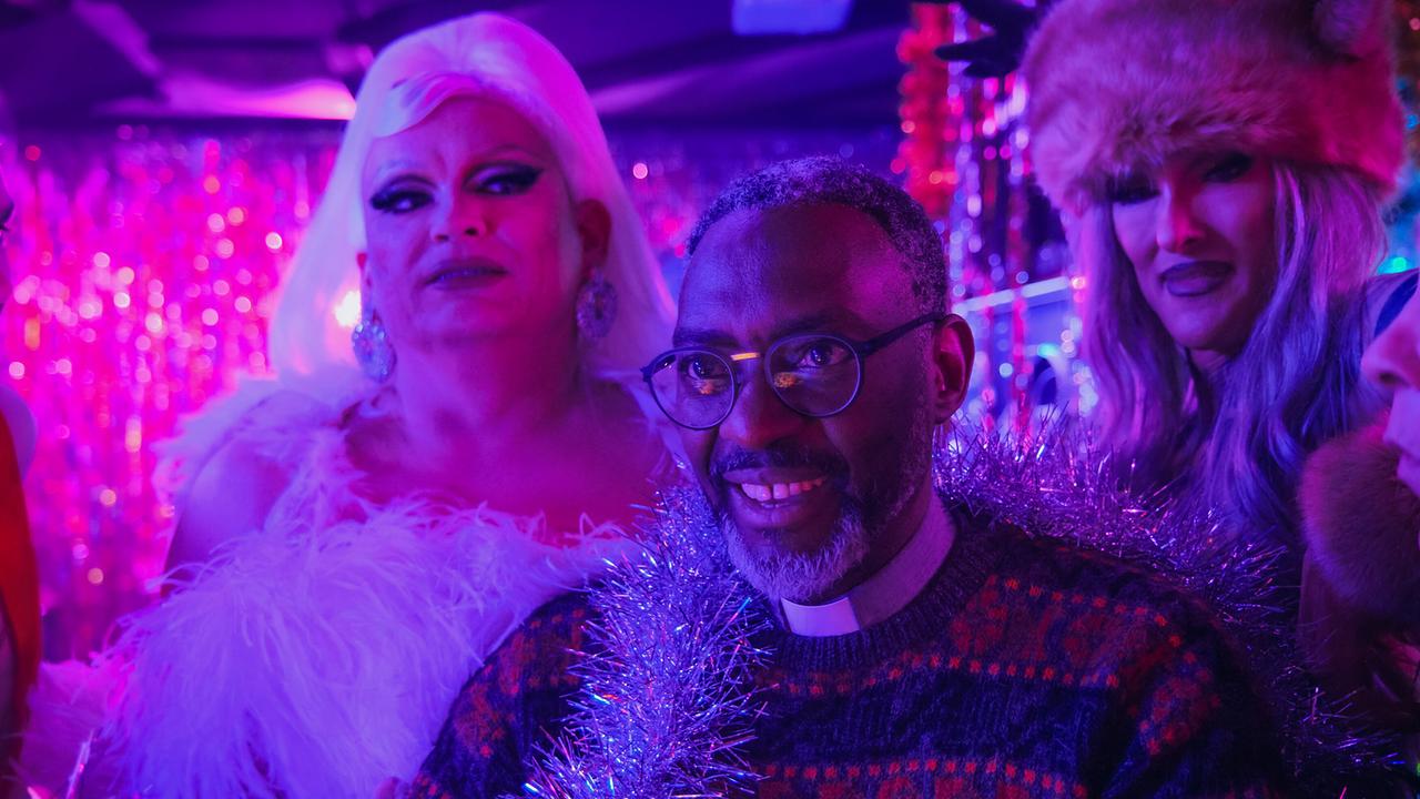 Pater Ramson steht mit Zola und Francine in einer farbenfroh beleuchteten Partyszene voller Deko und Glitzer.