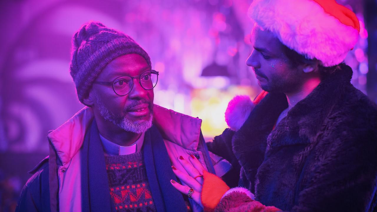 Tom mit Weihnachtsmütze legt Pater Ramson mit Haube und Anorak in einer lila ausgeleuchteten Umgebung die Hand auf die Schulter.