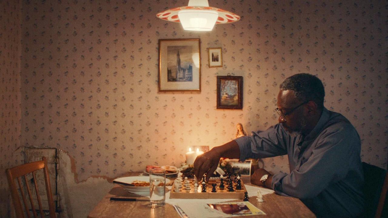 Pater Ramson sitzt an einem Esstisch in einem gemütlich beleuchteten, etwas altmodischen Raum und spielt mit sich konzentriert eine Partie Schach.