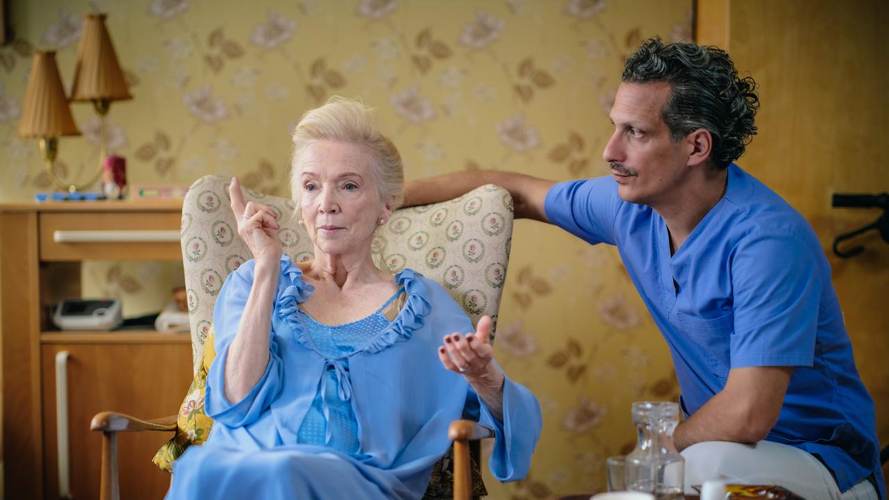 Mathilde Eisenhut sitzt in einem Sessel und spricht mit einer Pflegekraft in einem gemütlichen Wohnraum.