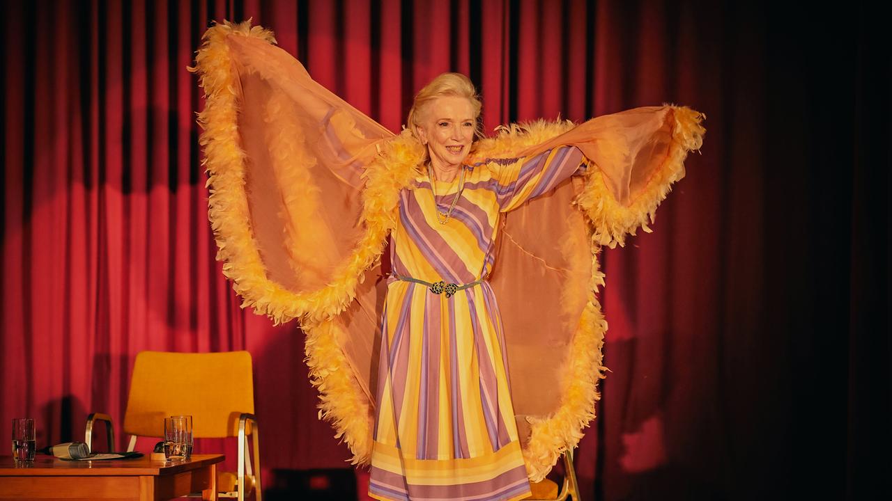 Mathilde Eisenhut im farbenfrohen Kleid steht auf der Bühne und breitet einen großen, federbesetzten Umhang aus.