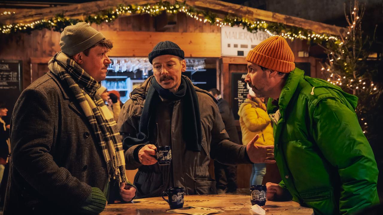 Roland, Christian und Max stehen an einem Stehtisch auf dem Weihnachtsmarkt und diskutieren bei warmen Getränken.