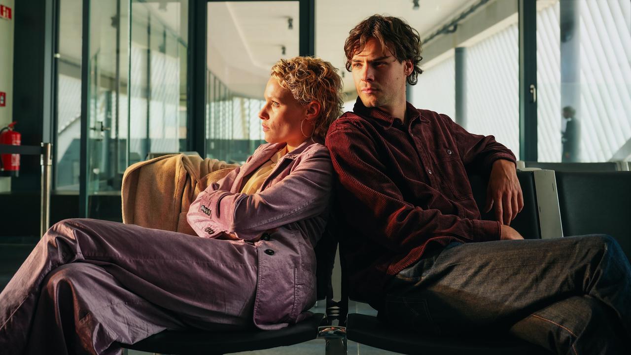 Silvia Holzbauer und Fabian Polly lehnen sich im Wartebereich eines Flughafengates entspannt aneinander.