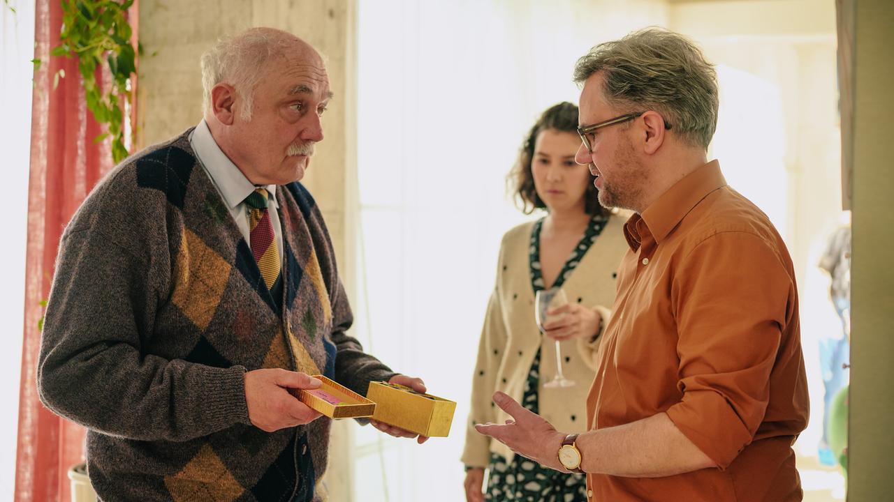 Herbert zeigt Richard eine geöffnete goldene Geschenkbox. Julia steht im Hintergrund.