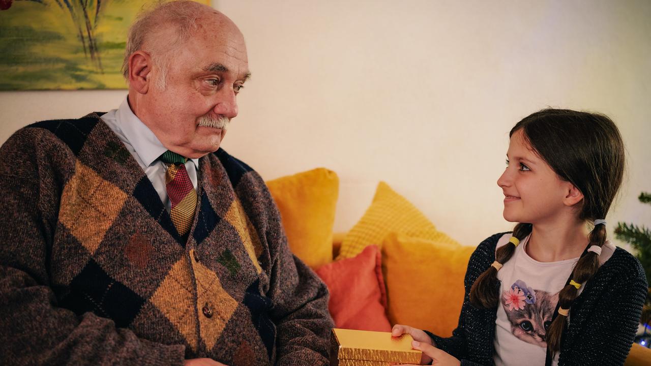 Herbert sitzt mit dem Mädchen Emily auf dem Sofa, die ihm eine goldene Geschenkbox reicht.
