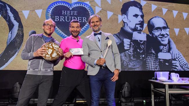 Dr. Florian Freistetter, Martin Puntigam und Univ.-Prof. Helmut Jungwirth stehen auf der "Science Busters"-Bühne  vor einem großen Hintergrundbild mit der Aufschrift „Hefe Brüder“; sie halten ein Brot, einen Umschlag und einen Kochlöffel.