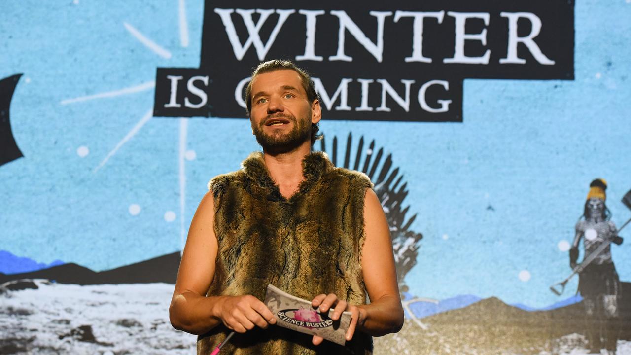 © ORF/Hubert Mican Martin Puntigam in einem ärmellosen Felloberteil steht auf der Bühne vor einer Leinwand mit der Aufschrift „Winter is Coming“.