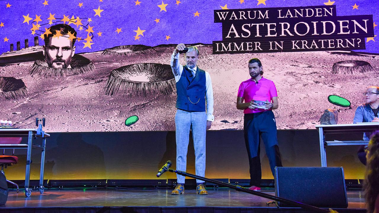 Univ.-Prof. Dr. Helmut Jungwirth, Martin Puntigam und Dr. Florian Freistetter auf der Bühne vor einer großen Projektionsfläche, auf der die Frage „Warum landen Asteroiden immer in Kratern?“ zu lesen ist.
