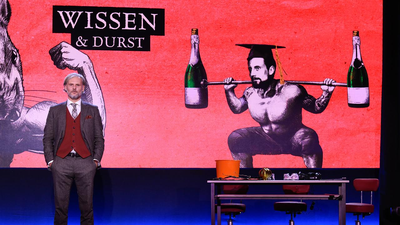 Univ.-Prof. Dr. Helmut Jungwirth steht auf der Bühne vor einer großen roten Grafik mit der Aufschrift „Wissen & Durst“, die eine stilisierte Figur mit Flaschen zeigt, daneben ein Tisch mit Requisiten.