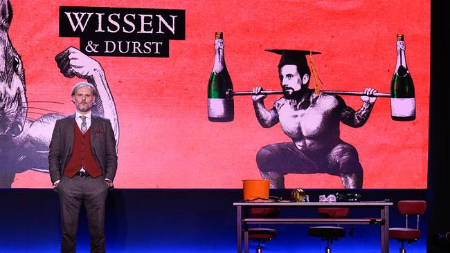 Univ.-Prof. Dr. Helmut Jungwirth steht auf der Bühne vor einer großen roten Grafik mit der Aufschrift „Wissen & Durst“, die eine stilisierte Figur mit Flaschen zeigt, daneben ein Tisch mit Requisiten.