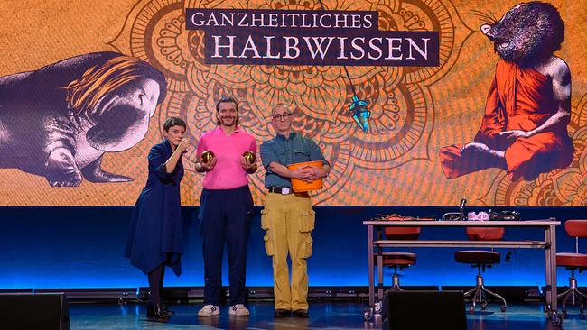 Dr. Elisabeth Oberzaucher, Martin Puntigam und "Gunkl" Günther Paal stehen auf einer Bühne vor einer großen Leinwand mit der Aufschrift „Ganzheitliches Halbwissen“, daneben stehen Requisiten und ein Tisch mit Technik.