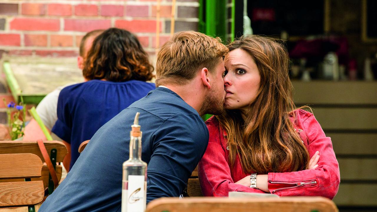 Marc und Teresa sitzen in einem Gastgarten an einem Tisch, er beugt sich zu ihr und küsst sie, sie erwidert den Kuss blickt jedoch jemanden erschrocken über seine Schulter an