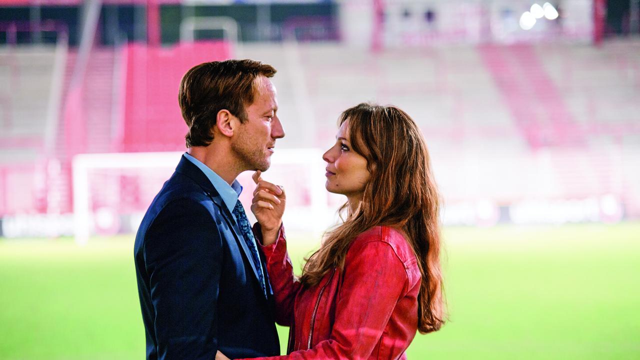 Wotan Wilke Möhring als Alex in blauem Anzug und  Mina Tander als Teresa in roter Lederjacke, in den Sitzrängen eines leeren Fußballstadions umarmen sie sich und blicken einander freundlich in die Augen