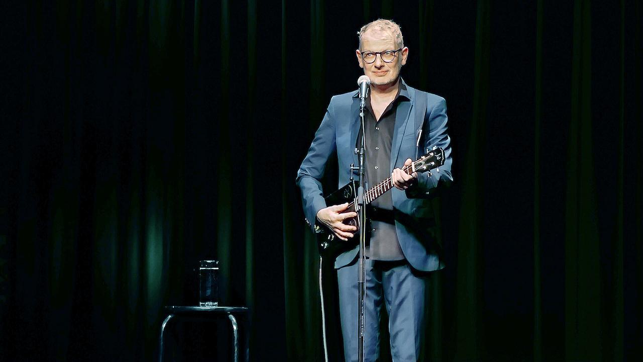 Severin Gröbner im blauen Anzug spielt auf einer dunklen Bühne Gitarre und steht dabei vor einem Mikrofon.