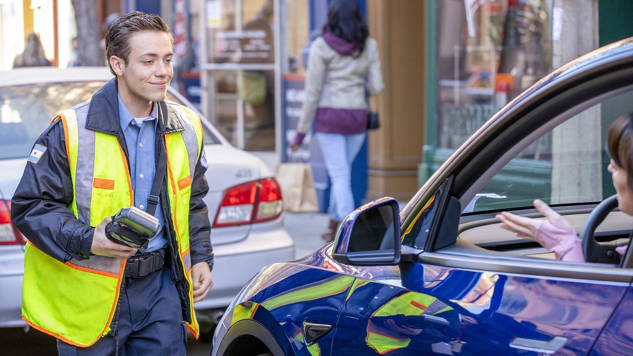 Carl Gallagher als Parkwächter auf der Straße, eine im Auto sitzende Frau spricht mit ihm, er lächelt sie freundlich an