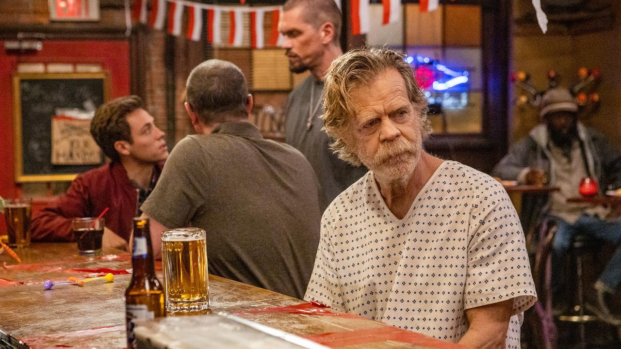 William H. Macy sitzt als Frank Gallagher an der Theke eines Pubs und trägt offensichtlich einen Krankenhauskittel