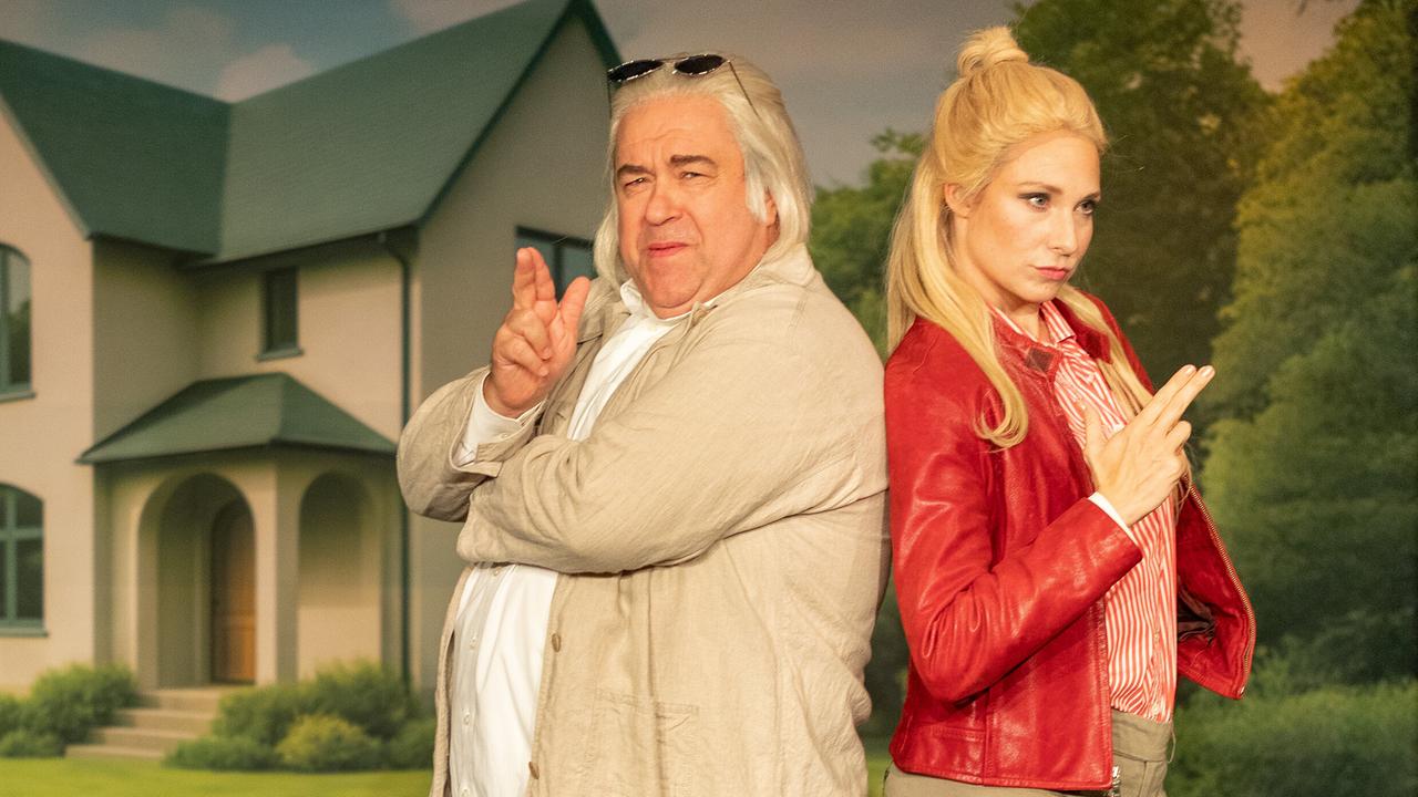 Bernhard Murg und Jenny Frankl in roter Lederjacke stehen vor einem Einfamilienhaus-Bühnenbild lässig Rücken an Rücken mit Fingerpistolen in einer überzeichneten Agentenpose und .