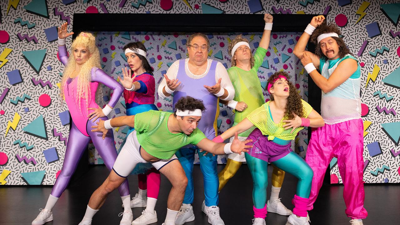 Jenny Frankl, Katharina Dorian, Matthias Mamedof, Bernhard Murg, Joachim Brandl, Ariana Schirasi-Fard und Julian Loidl posieren in bunten Trainingsoutfits vor einer bunten Kulisse wie bei einem Aerobic-Workout der 1980er-Jahre.