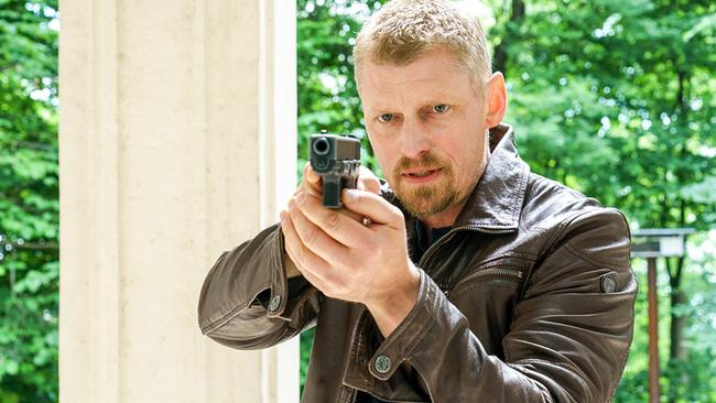 Martin Gruber als Max Herzog richtet seine Pistole auf jemanden im Grünen.