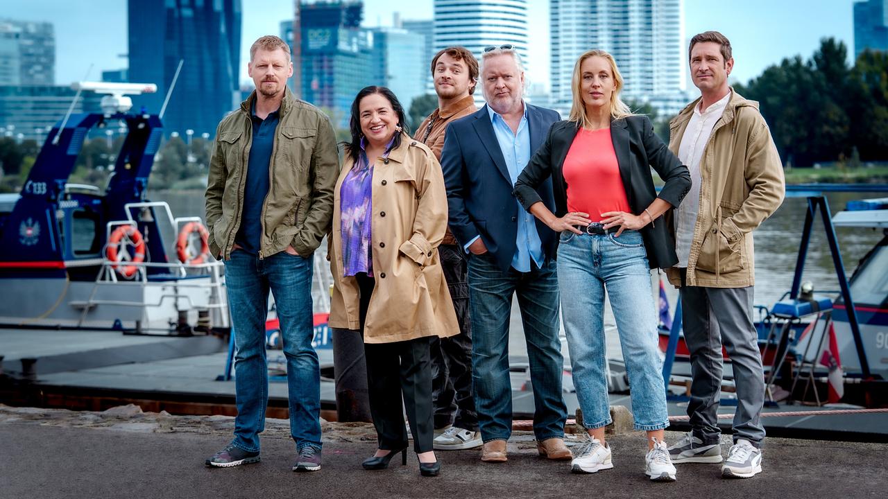 Martin Gruber, Maria Happel, Max Fischnaller, Gregor Seberg, Lilian Klebow und Andreas Kiendl stehen am Ufer vor einem Boot, im Hintergrund ist die Skyline von Wien zu sehen.