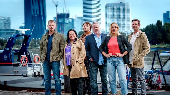 Martin Gruber, Maria Happel, Max Fischnaller, Gregor Seberg, Lilian Klebow und Andreas Kiendl stehen am Ufer vor einem Boot, im Hintergrund ist die Skyline von Wien zu sehen.
