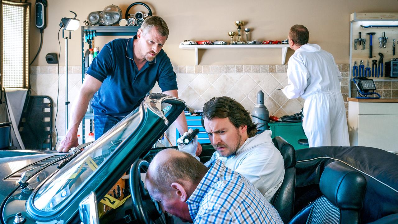 © ORF/Satel Film/Petro Domenigg In einer Garage untersuchen Max Herzog und Julius Rubatsch den Toten im Cabrio, während im Hintergrund ein weiterer Ermittler arbeitet.