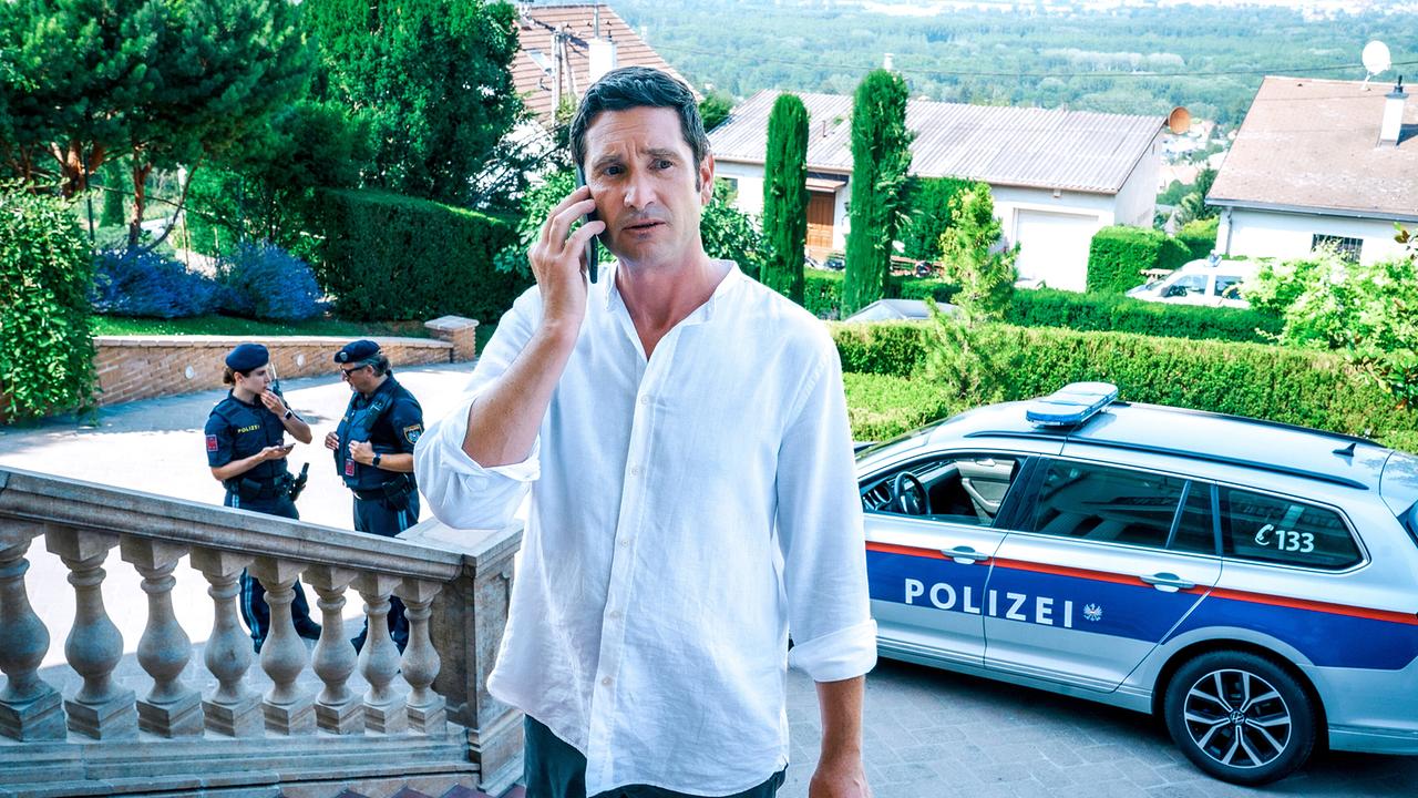 © ORF/Satel Film/Petro Domenigg Klaus Lechner telefoniert auf einer Außentreppe mit Balustrade, im Hintergrund stehen Polizeibeamte neben einem Polizeiauto.