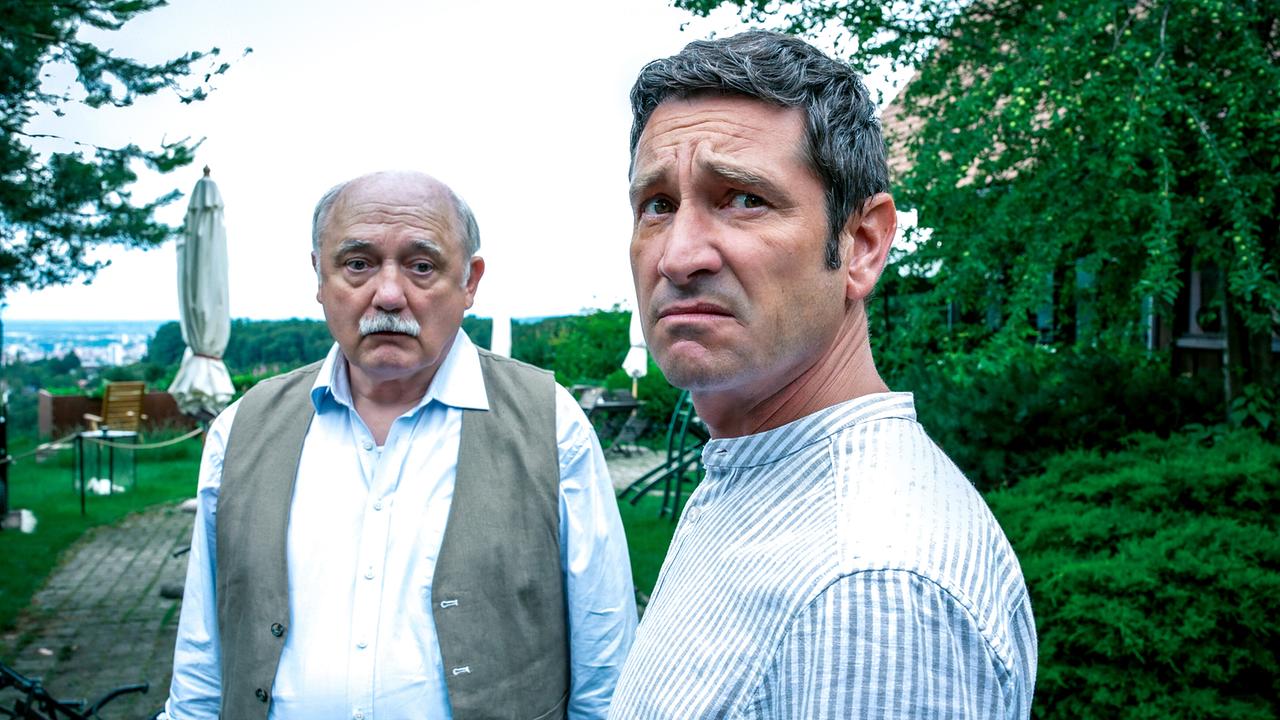 Draußen im Garten stehen Erwin Happich und Klaus Lechner bei bewölktem Wetter beisammen, im Hintergrund sind eine Terrasse und Landschaft zu sehen.