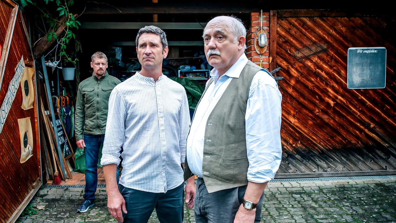 Vor einer Holzscheune stehen Klaus Lechner und Erwin Happich, während Max Herzog im Hintergrund am Eingang steht.