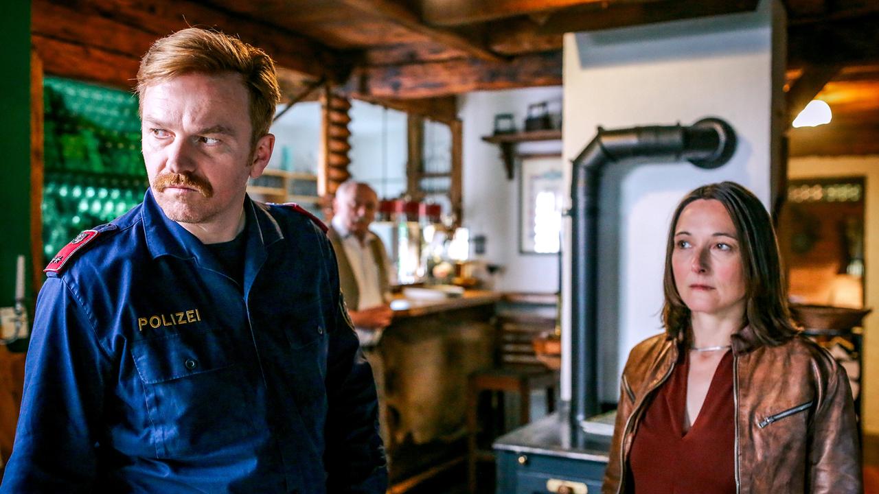 In einem rustikalen Gastraum stehen Polizist Sonny Raimann und Ellen Raimann vor einem alten Ofen und schauen aufmerksam in den Raum.