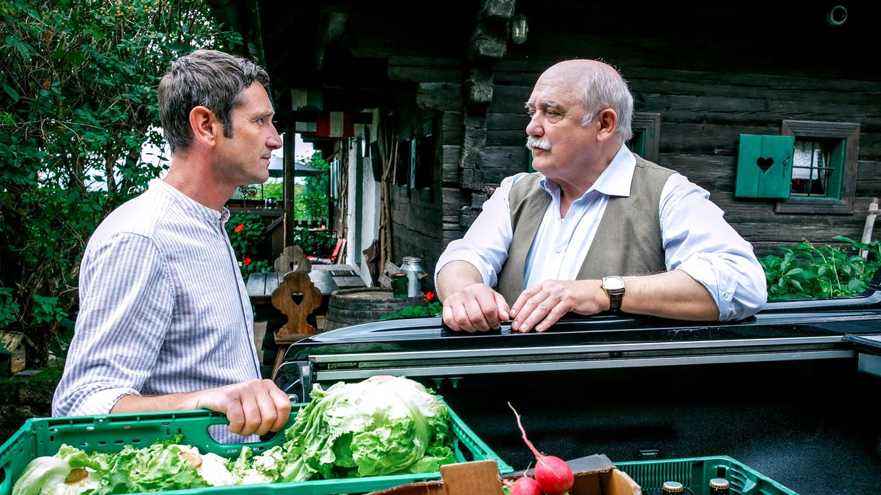 Neben einem Wagen mit Gemüsekisten unterhalten sich Klaus Lechner und Erwin Happich vor einem ländlichen Holzhaus.