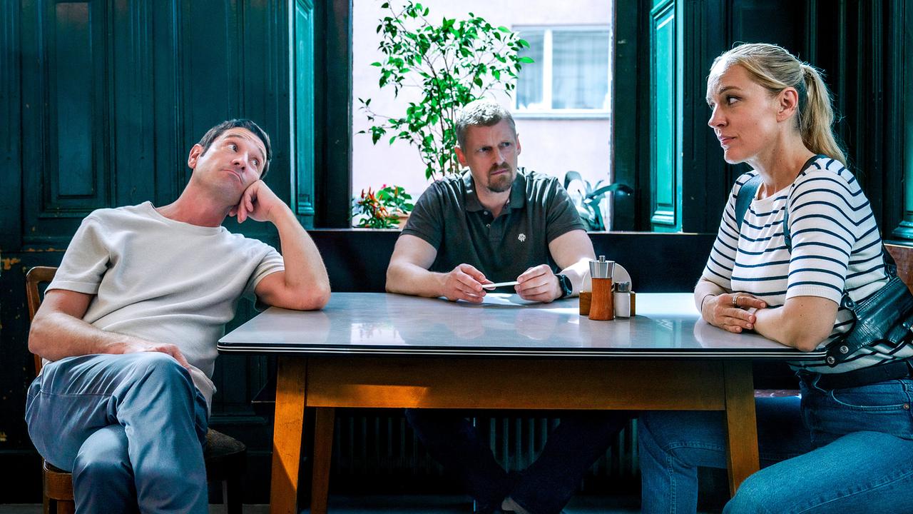 Klaus Lechner, Max Herzog und Penny Lanz sitzen an einem Tisch in einem Wirtshaus und wirken nachdenklich.