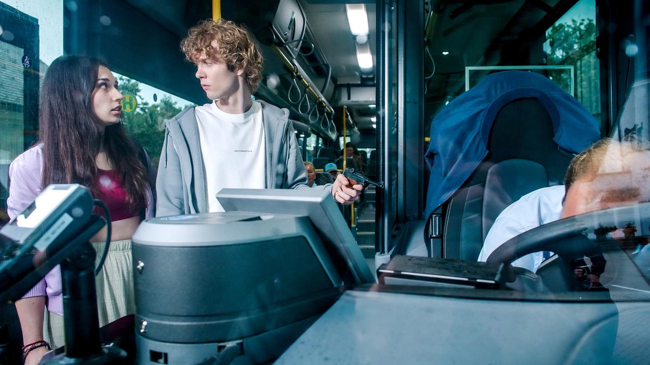 Patricia Kilb und Manuel Wallner, der eine Pistole in der Hand hält, stehen im Eingangsbereich eines Busses, am Fahrersitz liegt der tote Busfahrer mit Kopf auf dem Lenkrad.