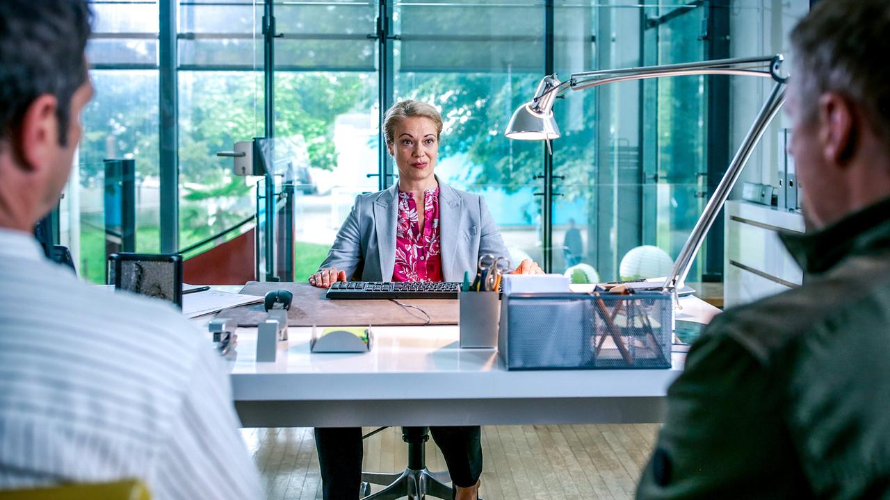 Maria Geyer sitzt an einem Schreibtisch in einem hellen, verglasten Büro mit Computer und Arbeitsutensilien mit Blick ins Grüne. Ihr gegenüber sitzen Klaus Lechner und Max Herzog, die im Vordergrund von hinten zu sehen sind. 
