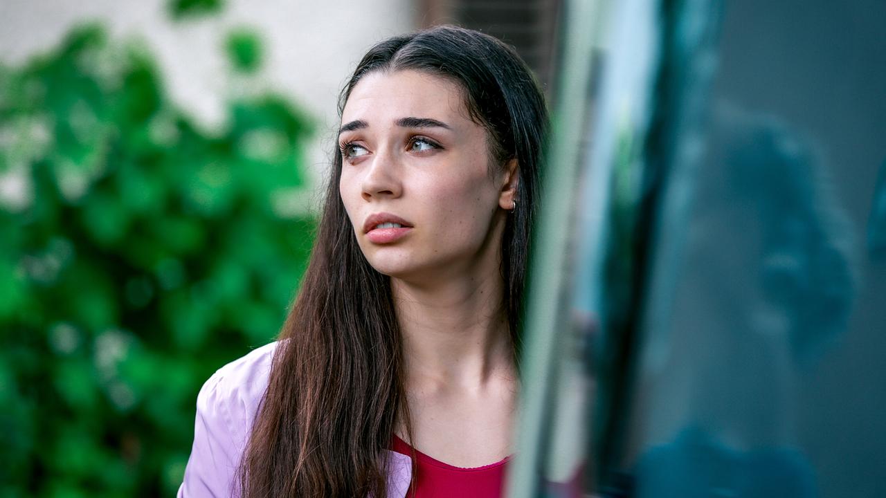 Patricia Kilb steht im Freien teilweise hinter einem Objekt, umgeben von Grün und Gebäuden.