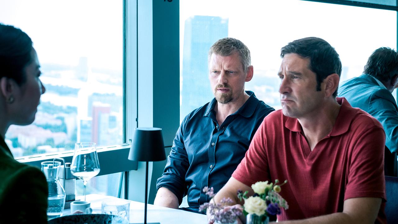 Max Herzog und Klaus Lechner sitzen gegenüber von Bao Chen an einem gedeckten Tisch in einem modernen Restaurant mit Blick auf die Stadt.