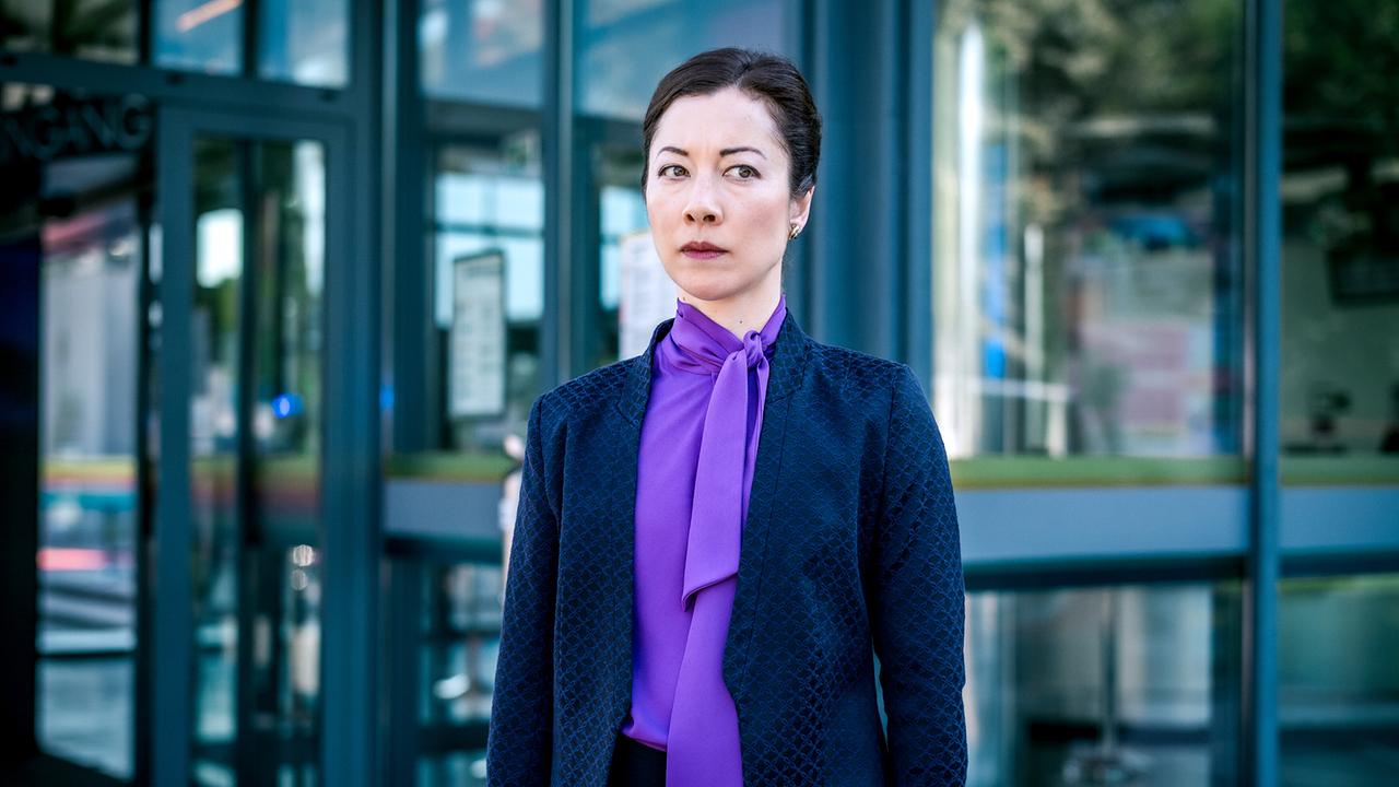 Bao Chen in eleganter Businesskleidung steht vor einem gläsernen Eingangsbereich eines modernen Gebäudes.