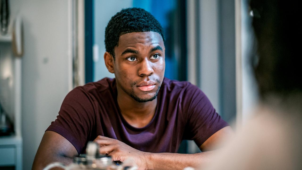 Hamza Biobaku sitzt an einem Tisch in einem Innenraum Ayo Ajiri, die unscharf im Vordergrund zu sehen ist, gegenüber.